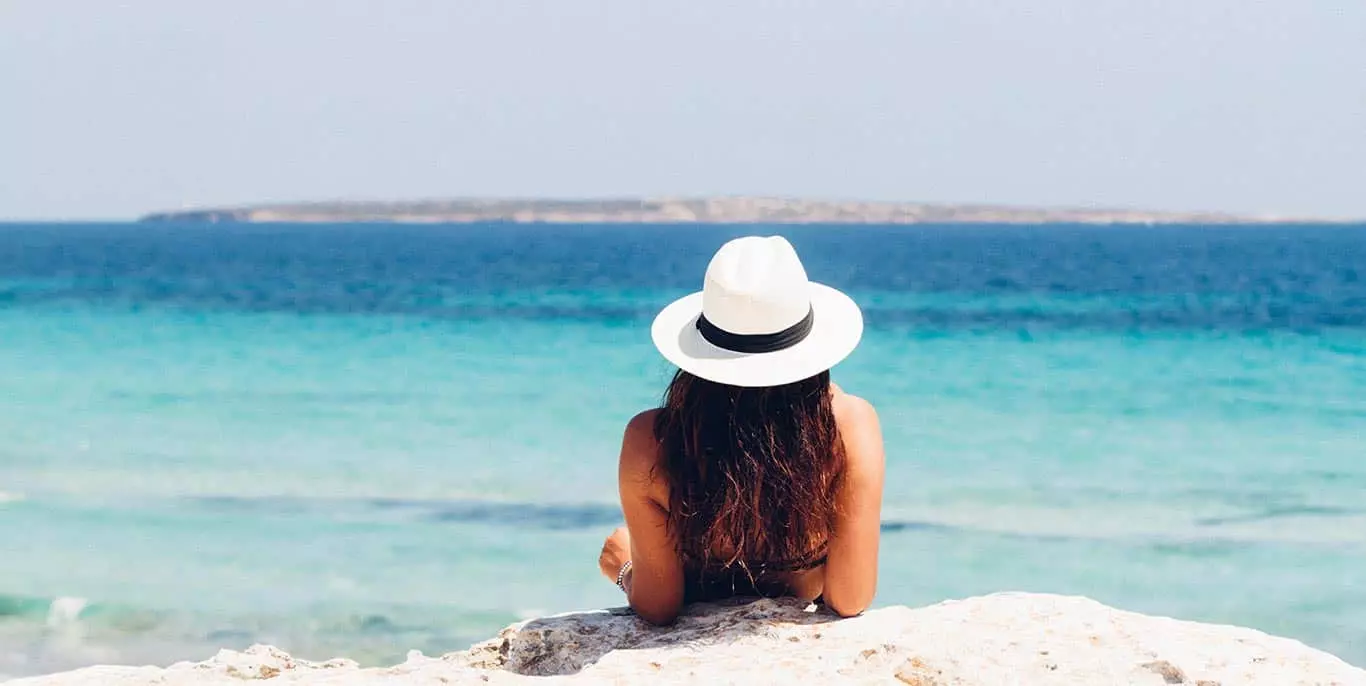 Rückansicht einer Frau mit Hut in Strandbekleidung an Steinwand lehnend mit Blick auf blauem Meer mit Insel am Horizont
